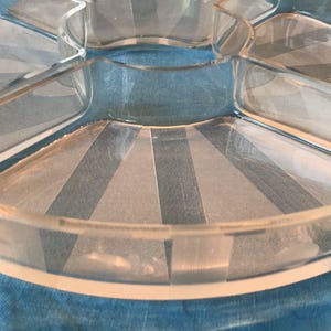 Peut inclure: Plat de service en verre transparent à cinq compartiments. Le plat a une forme circulaire avec une zone centrale ronde et quatre sections en forme de tarte. Le verre présente un motif subtil de rayures verticales. Le plat est posé sur une surface bleue.