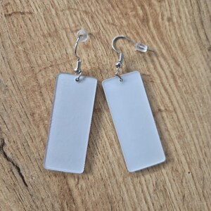 May include: A pair of rectangular, frosted white earrings with silver-colored fish hook ear wires. The earrings are displayed on a wooden surface.