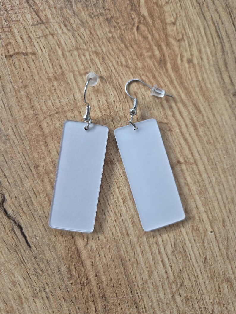 May include: A pair of rectangular, frosted white earrings with silver-colored fish hook ear wires. The earrings are displayed on a wooden surface.