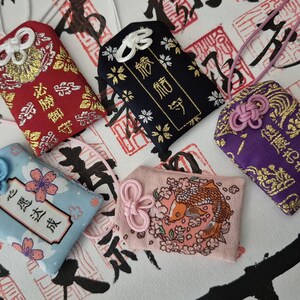 May include: A collection of Japanese omamori charms in various colors. The charms feature intricate designs, including floral patterns, koi fish, and kanji characters. Each charm has a decorative knot and a cord for carrying.