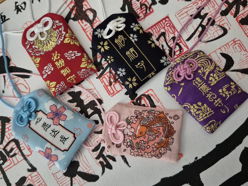 May include: A collection of Japanese omamori charms in various colors. The charms feature intricate designs, including floral patterns, koi fish, and kanji characters. Each charm has a decorative knot and a cord for carrying.