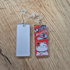 May include: A pair of rectangular earrings. One earring is a solid white rectangle. The other earring has a red background with Japanese text, koi fish, and a cartoon cat with its paws together.