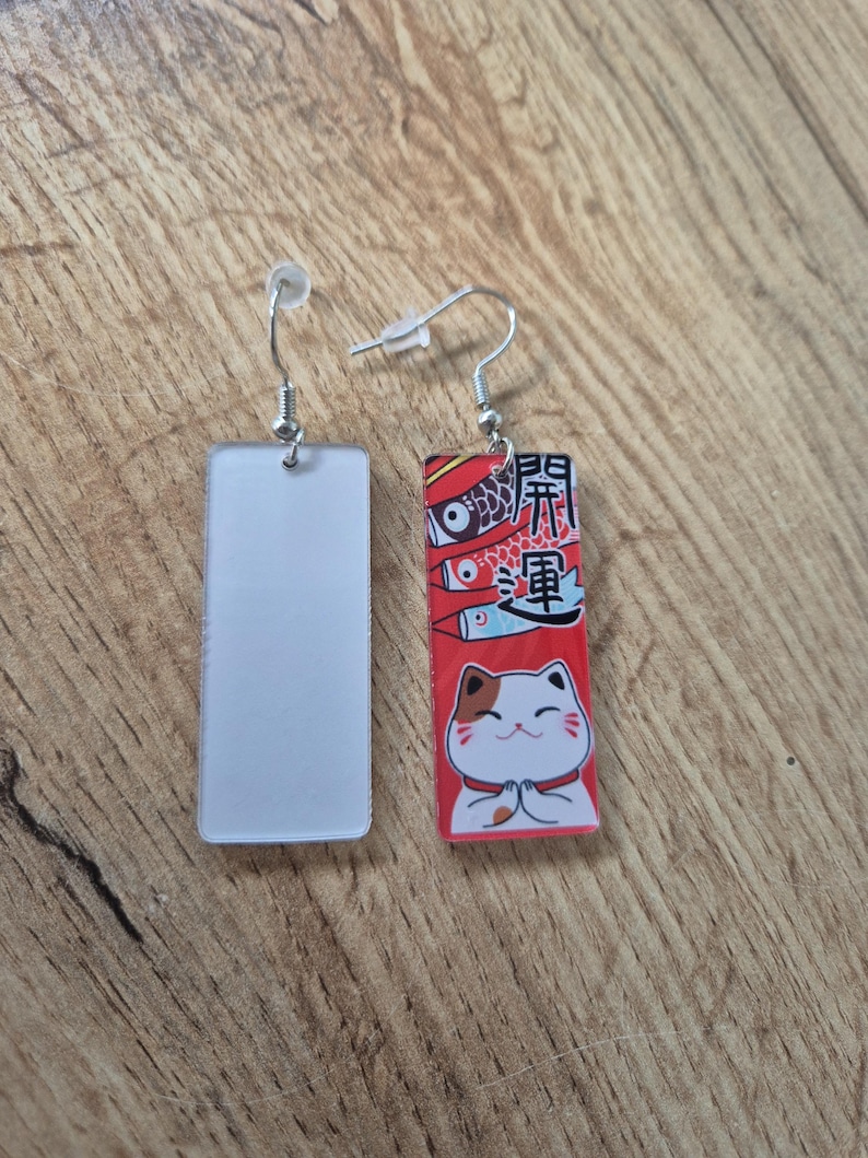 May include: A pair of rectangular earrings. One earring is a solid white rectangle. The other earring has a red background with Japanese text, koi fish, and a cartoon cat with its paws together.