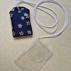 May include: A navy blue fabric charm with white and gold floral patterns, attached to a white cord. A clear plastic card holder is also present, connected to the same cord. The items are on a light tan surface.