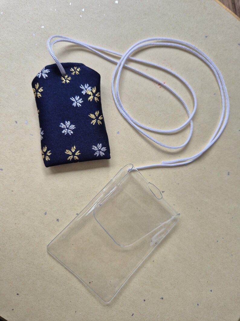 May include: A navy blue fabric charm with white and gold floral patterns, attached to a white cord. A clear plastic card holder is also present, connected to the same cord. The items are on a light tan surface.