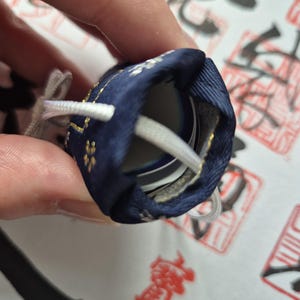 May include: A navy blue drawstring pouch with gold floral accents and white drawstrings. The pouch is partially open, revealing a glimpse of the contents inside. The background features Japanese calligraphy.