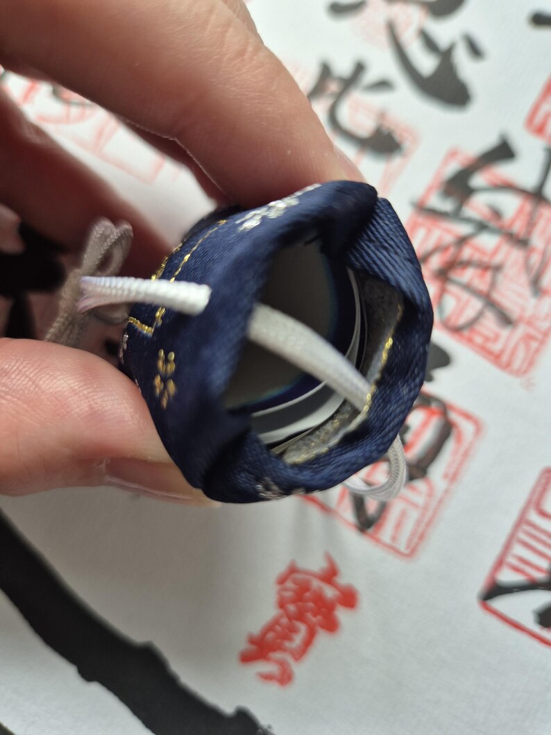 May include: A navy blue drawstring pouch with gold floral accents and white drawstrings. The pouch is partially open, revealing a glimpse of the contents inside. The background features Japanese calligraphy.