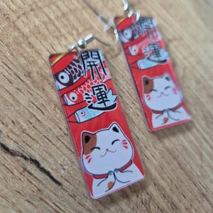 May include: A pair of rectangular earrings with silver hooks. Each earring features a red background with Japanese text, a cartoon cat, and koi fish illustrations. The cat has white fur, brown markings, and is in a prayerful pose.