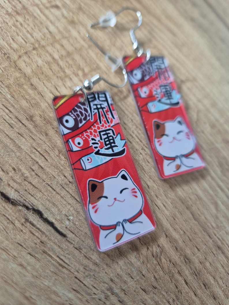 May include: A pair of rectangular earrings with silver hooks. Each earring features a red background with Japanese text, a cartoon cat, and koi fish illustrations. The cat has white fur, brown markings, and is in a prayerful pose.