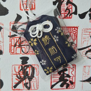 May include: A navy blue Japanese omamori charm with gold floral accents and gold text, encased in clear plastic. The charm is tied with a white cord and bow. The background features red and black Japanese calligraphy stamps.