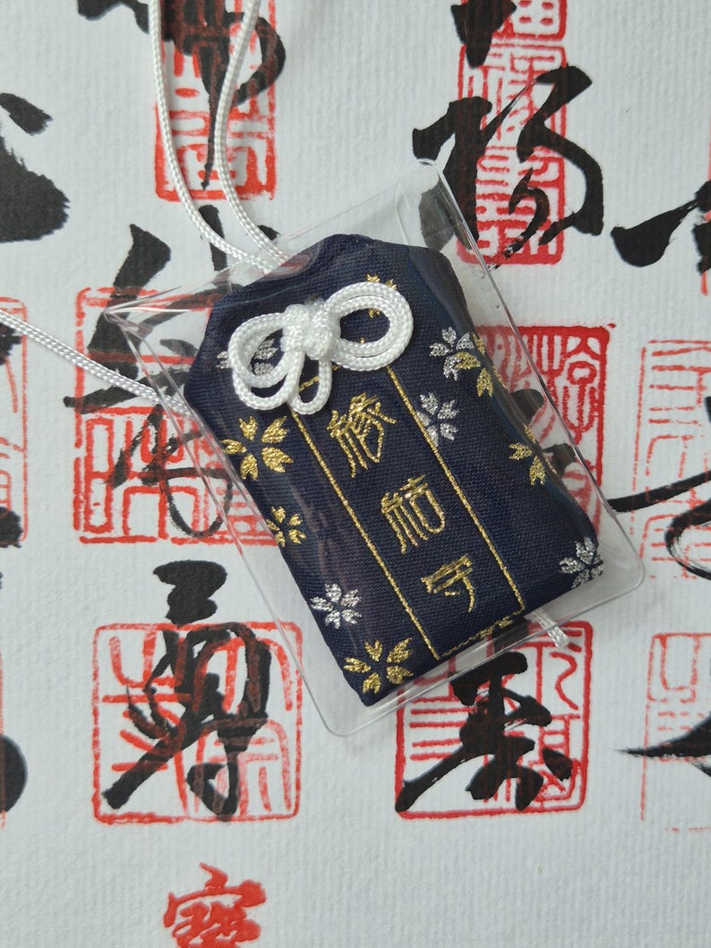May include: A navy blue Japanese omamori charm with gold floral accents and gold text, encased in clear plastic. The charm is tied with a white cord and bow. The background features red and black Japanese calligraphy stamps.