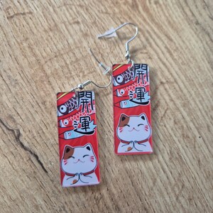 May include: A pair of rectangular earrings featuring a red background with a Japanese lucky cat design. The earrings have a white cat with brown spots, fish, and Japanese text. The earrings hang from silver-colored hooks.
