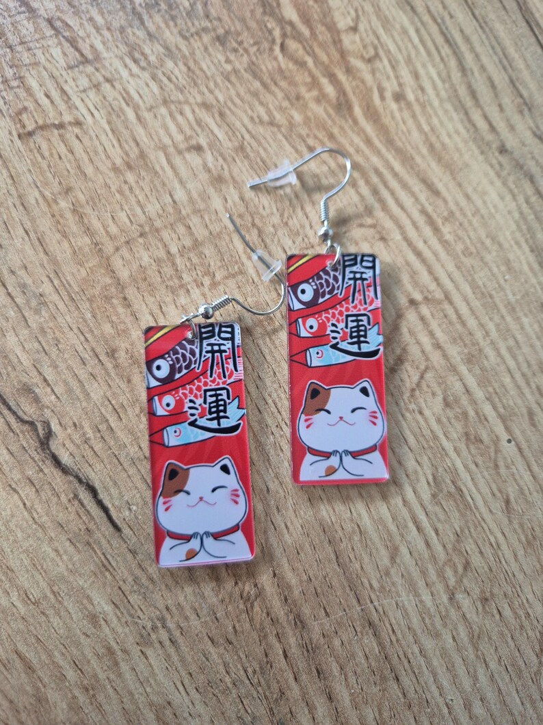 May include: A pair of rectangular earrings featuring a red background with a Japanese lucky cat design. The earrings have a white cat with brown spots, fish, and Japanese text. The earrings hang from silver-colored hooks.