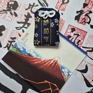 May include: A navy blue Japanese omamori charm with gold text and floral accents, hanging from a white cord. The charm rests on a postcard featuring a mountain landscape with a red peak and blue sky.