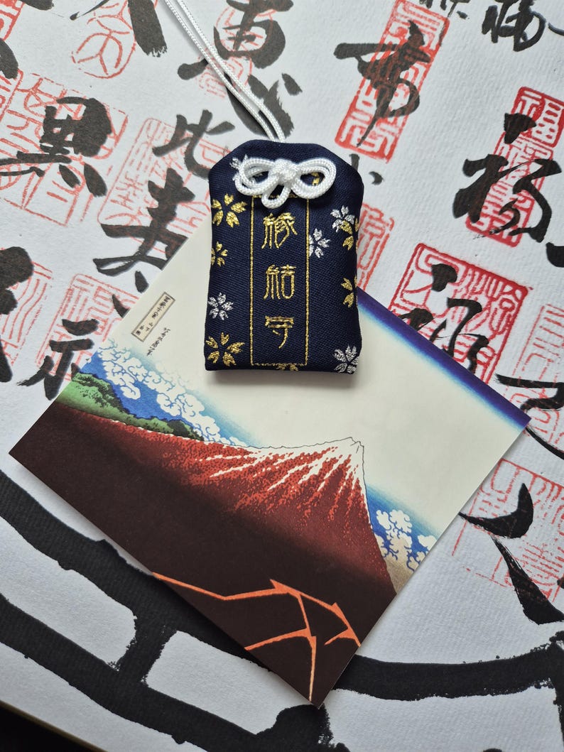 May include: A navy blue Japanese omamori charm with gold text and floral accents, hanging from a white cord. The charm rests on a postcard featuring a mountain landscape with a red peak and blue sky.
