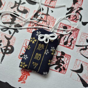 May include: A navy blue Japanese omamori charm with gold and silver floral accents and Japanese text. The charm is attached to a white cord and sits on a white surface with red and black calligraphy.