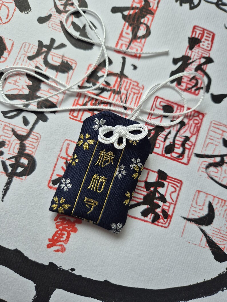 May include: A navy blue Japanese omamori charm with gold and silver floral accents and Japanese text. The charm is attached to a white cord and sits on a white surface with red and black calligraphy.