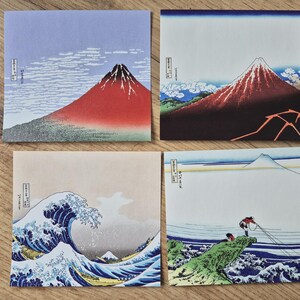 May include: Four square art prints featuring Japanese landscape scenes. The prints depict Mount Fuji, a large ocean wave, and figures fishing. Each print has a unique color palette, including red, blue, and beige.