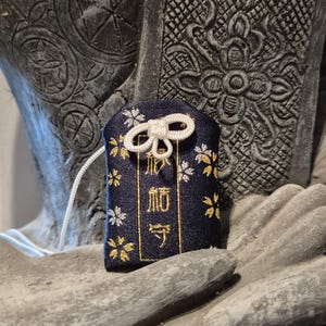 May include: A navy blue Japanese omamori charm with gold and white floral accents and Japanese text. The charm has a white bow and a white string. The charm is resting on a gray stone surface.