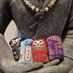 May include: Four colorful Japanese omamori charms with intricate designs and text, held in a stone sculpture's hand. The charms are navy, light blue, red, and purple, each with a unique knot and cord. A beaded necklace with a lotus pendant is also visible.