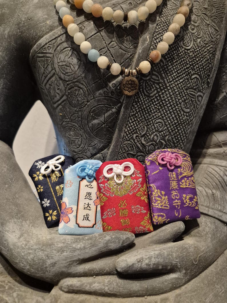 May include: Four colorful Japanese omamori charms with intricate designs and text, held in a stone sculpture's hand. The charms are navy, light blue, red, and purple, each with a unique knot and cord. A beaded necklace with a lotus pendant is also visible.