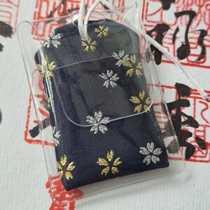 May include: A navy blue fabric charm with gold and silver floral designs, encased in a clear plastic sleeve. A white string is attached for hanging. The background features red and black Japanese calligraphy.