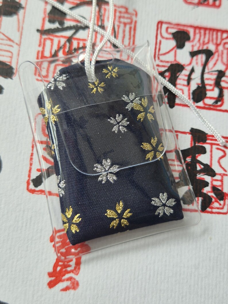 May include: A navy blue fabric charm with gold and silver floral designs, encased in a clear plastic sleeve. A white string is attached for hanging. The background features red and black Japanese calligraphy.