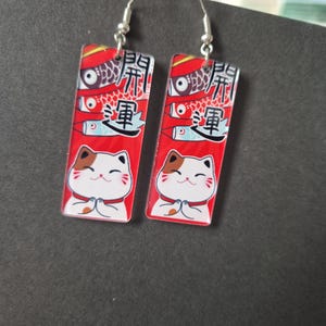 May include: A pair of rectangular earrings with silver hooks. Each earring features a red background with Japanese text, a fish design, and a cartoon cat with its paws together. The cat has brown and white markings.