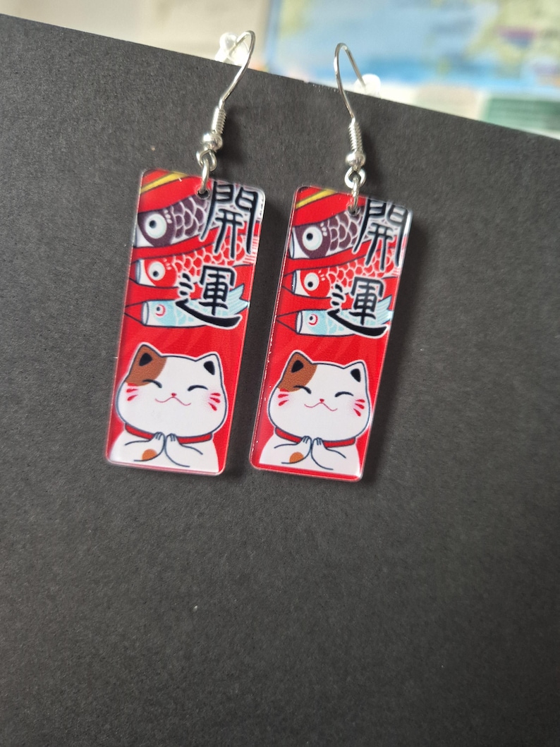 May include: A pair of rectangular earrings with silver hooks. Each earring features a red background with Japanese text, a fish design, and a cartoon cat with its paws together. The cat has brown and white markings.