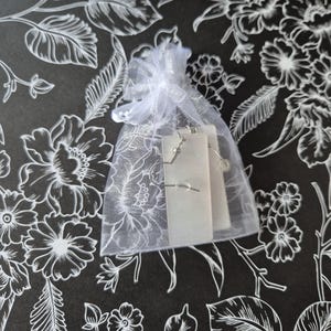 May include: A small, white, sheer gift bag tied with a ribbon, containing a pair of silver earrings on a white card. The background is a black surface with a white floral pattern.