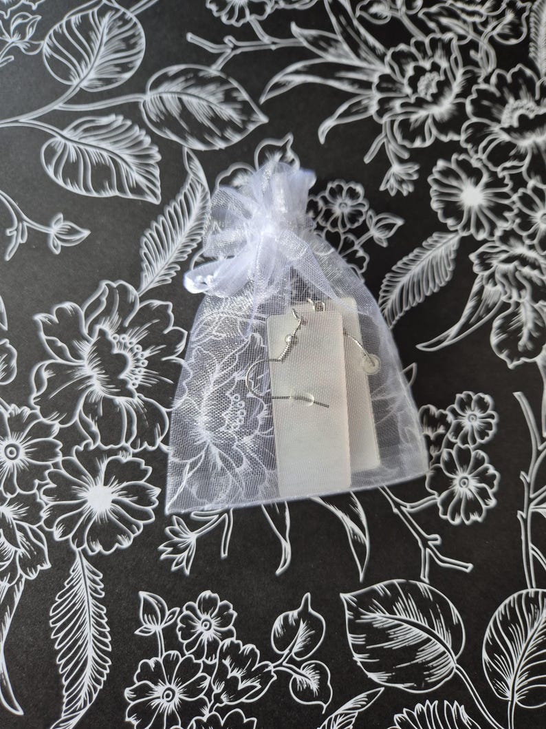 May include: A small, white, sheer gift bag tied with a ribbon, containing a pair of silver earrings on a white card. The background is a black surface with a white floral pattern.