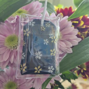 May include: A dark blue item with gold and silver floral designs is encased in a clear plastic holder, suspended by a white string. The item is set against a backdrop of pink and red flowers and green leaves.