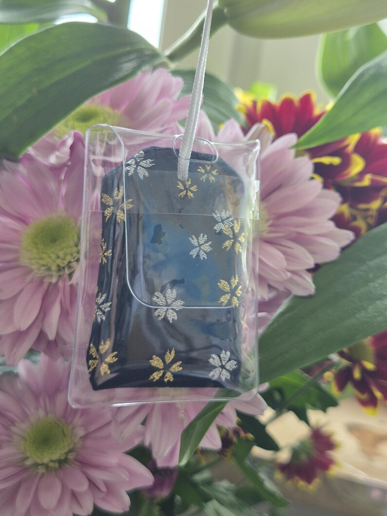 May include: A dark blue item with gold and silver floral designs is encased in a clear plastic holder, suspended by a white string. The item is set against a backdrop of pink and red flowers and green leaves.