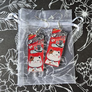 May include: A pair of red and white Maneki Neko cat earrings. Each earring features a smiling cat with its paws together, and Japanese text. The earrings are displayed in a small, sheer white gift bag against a black floral background.