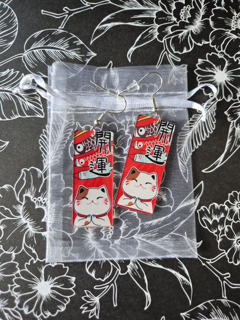 May include: A pair of red and white Maneki Neko cat earrings. Each earring features a smiling cat with its paws together, and Japanese text. The earrings are displayed in a small, sheer white gift bag against a black floral background.