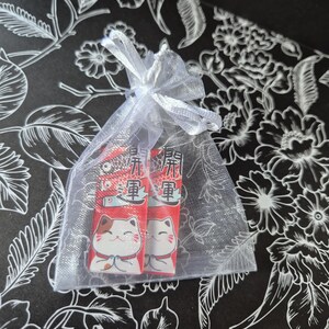 May include: Two red and white Japanese lucky cat charms inside a sheer white organza gift bag tied with a white ribbon. The charms have Japanese text and a smiling cat design. The background is black with a white floral pattern.
