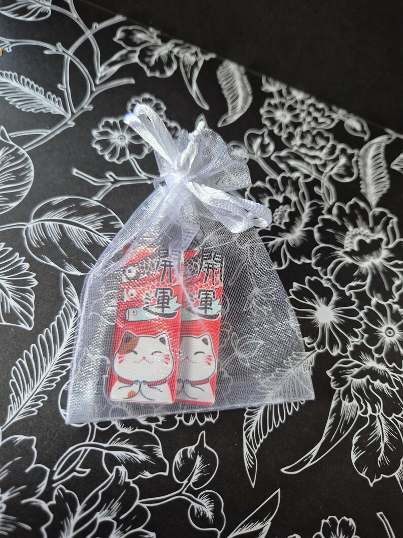 May include: Two red and white Japanese lucky cat charms inside a sheer white organza gift bag tied with a white ribbon. The charms have Japanese text and a smiling cat design. The background is black with a white floral pattern.