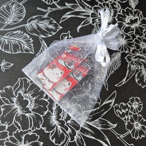 May include: Two red and white Japanese lucky cat charms inside a sheer white organza bag tied with a white ribbon. The background is black with a white floral pattern.