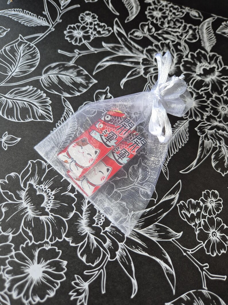 May include: Two red and white Japanese lucky cat charms inside a sheer white organza bag tied with a white ribbon. The background is black with a white floral pattern.