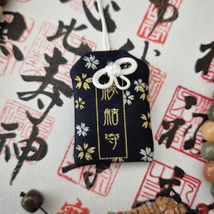 May include: A navy blue Japanese omamori charm with gold text and white and gold floral accents. The charm is attached to a white cord with a decorative knot. The background features Japanese calligraphy and red stamps.