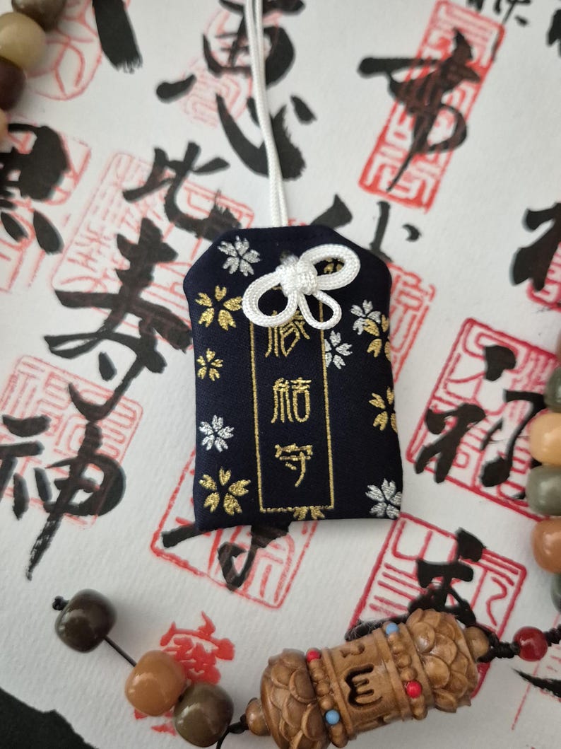 May include: A navy blue Japanese omamori charm with gold text and white and gold floral accents. The charm is attached to a white cord with a decorative knot. The background features Japanese calligraphy and red stamps.