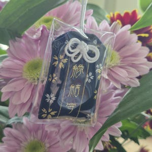 May include: A navy blue Japanese omamori charm with gold and white accents, hanging in front of a bouquet of pink and red flowers. The charm has Japanese characters and a white cord.