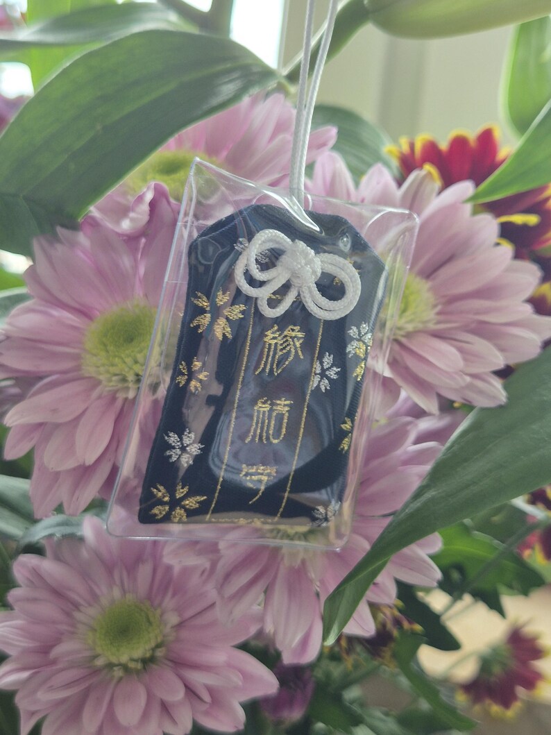 May include: A navy blue Japanese omamori charm with gold and white accents, hanging in front of a bouquet of pink and red flowers. The charm has Japanese characters and a white cord.