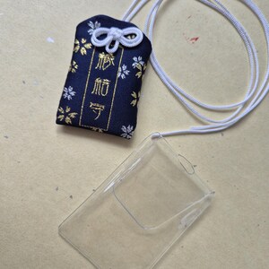 May include: A navy blue Japanese omamori charm with gold floral accents and white cord. The charm has Japanese text and is next to a clear plastic card holder.