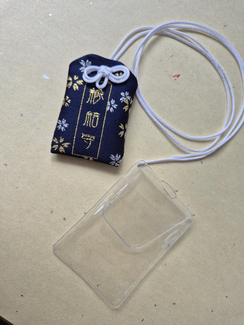 May include: A navy blue Japanese omamori charm with gold floral accents and white cord. The charm has Japanese text and is next to a clear plastic card holder.