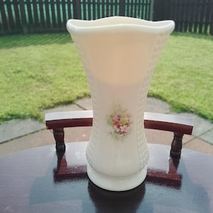 Puede incluir: Jarrón de cerámica color crema con un patrón texturizado similar a una cesta. El jarrón tiene una parte superior acampanada y un pequeño diseño floral en la parte delantera. Está sobre una mesa redonda de madera oscura. El jarrón mide unos 20 cm de alto.