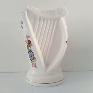 May include: White ceramic harp-shaped vase with a gold rim and floral accents. The vase features a detailed harp design with vertical lines and is decorated with red, blue, and pink flowers. A decorative piece for home decor.