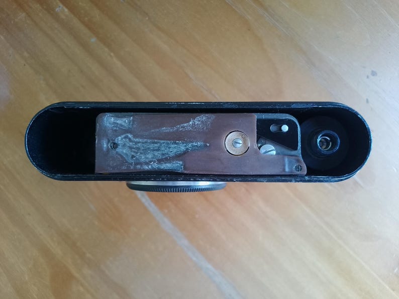 K&ouml;nnte beinhalten: Schwarzer Vintage-Kamerak&ouml;rper mit kupferfarbenem Innenmechanismus. Die Kamera hat einen Objektivanschluss und einen Filmtransportmechanismus. Die Oberseite der Kamera ist schwarz und abgerundet, mit einer strukturierten Oberfl&auml;che.