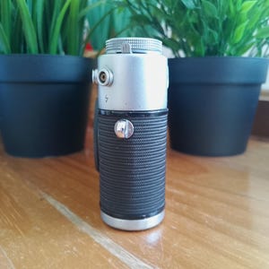 K&ouml;nnte beinhalten: Ein Objektiv einer Vintage-Kamera mit silbernem Ober- und Unterteil und einem schwarzen, strukturierten Griff. Das Objektiv hat einen kleinen silbernen Knopf und ein silbernes Zifferblatt. Der Hintergrund zeigt zwei gr&uuml;ne Topfpflanzen auf einer Holzoberfl&auml;che.