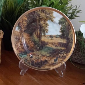 Könnte beinhalten: Dekorative Platte mit einer Landschaftsszene, die Bäume, einen Fluss und Figuren zeigt. Die Platte hat einen goldenen Rand und wird auf einem transparenten Kunststoffständer präsentiert. Das Kunstwerk zeigt eine pastorale Umgebung mit einer warmen Farbpalette.